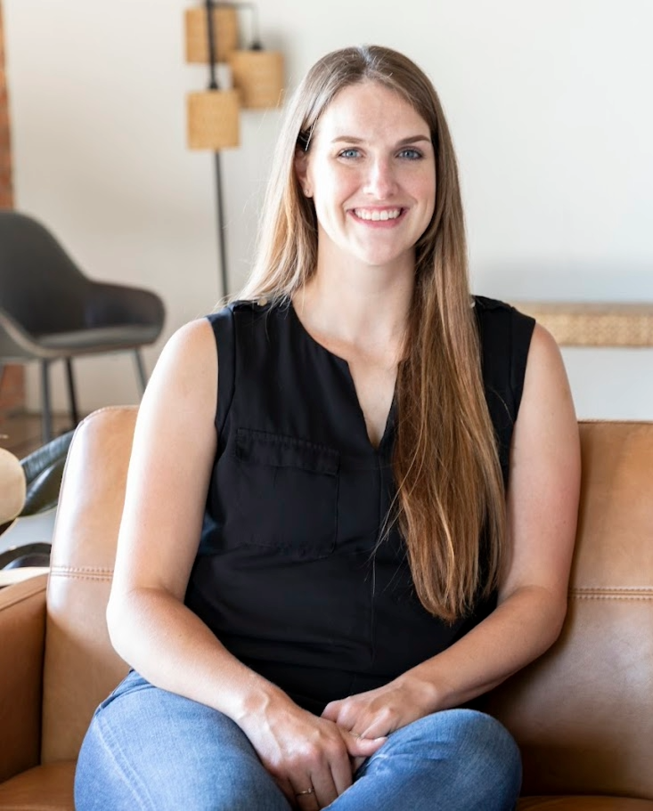 Smiling woman with long brown hair sits on a tan leather couch wearing a black sleeveless top and jeans in a bright room.