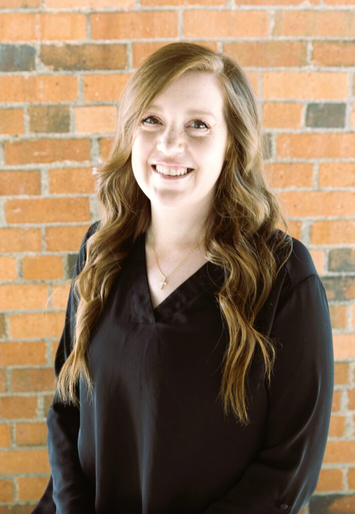 Smiling woman with long wavy light brown hair wearing a black top standing in front of a brick wall