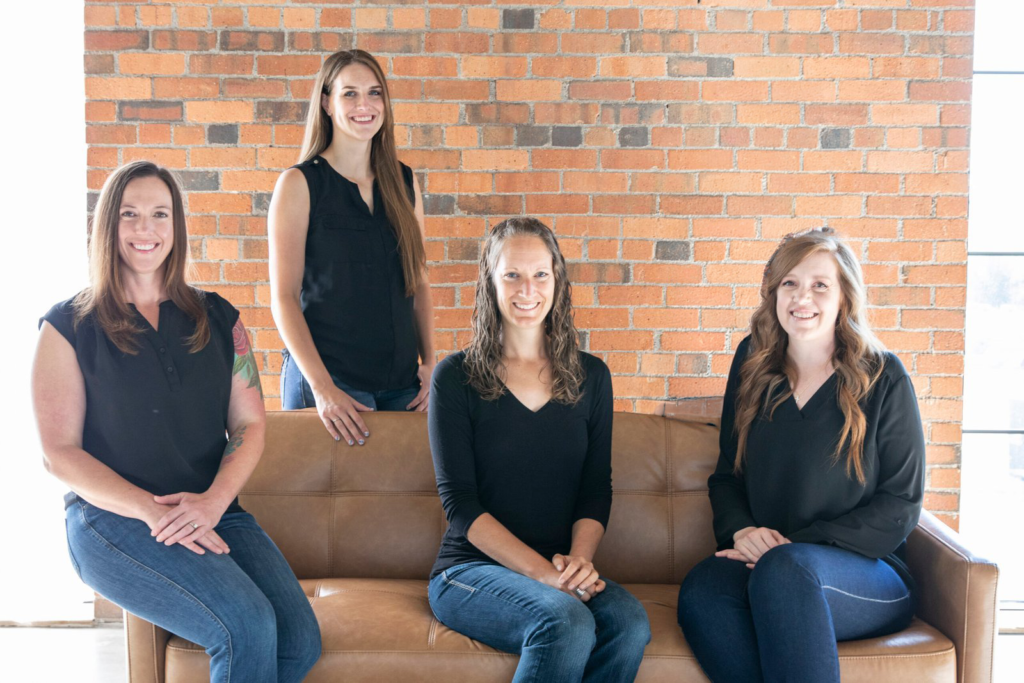 Four women sit and stand around a tan sofa in front of an orange brick wall, smiling at the camera.