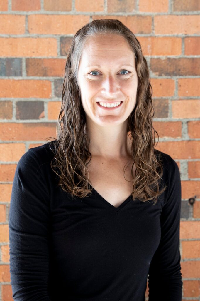 Smiling woman with wavy brown hair wearing a black V-neck top, standing in front of an orange brick wall.